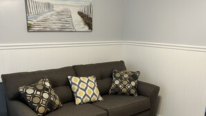 Un sofá gris oscuro con cojines estampados frente a una pared gris clara, con un cuadro con motivos playeros en la parte superior.
