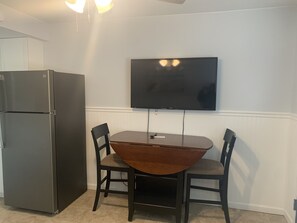 A compact kitchen with a built-in refrigerator, a wooden dining table with chairs, and a mounted flat-screen TV on the wall.
