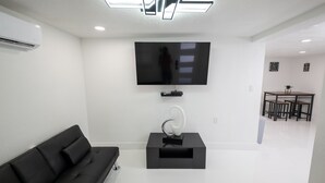 Una sala de estar moderna con un sofá negro, un televisor de pantalla plana montado en la pared, una mesita auxiliar con un objeto decorativo y un área de comedor al fondo.