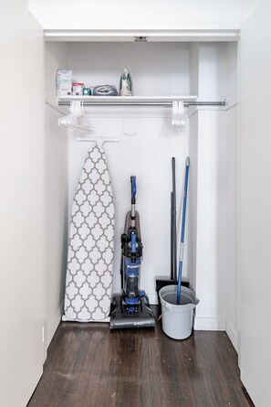 Un petit placard avec une tringle à vêtements, une planche à repasser, un aspirateur et un seau à serpillière.