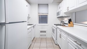 Una cocina compacta con armarios blancos, horno empotrado, microondas y una ventana con persianas.