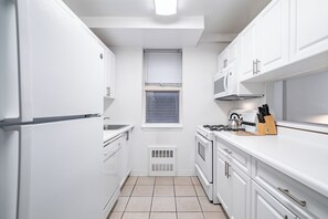 Une cuisine compacte avec des meubles blancs, un four encastré, un four à micro-ondes et une fenêtre équipée de stores.