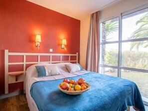 A hotel room with a bed, a bedside table, a fruit bowl on the bed, and a window with curtains. - Le Village (C4 010) by Interhome (Agay Village Cap Esterel)