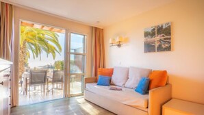 A modern living room with a sofa, a dining area, and a sliding glass door leading to an outdoor space with a palm tree. - Le Village (C4 010) by Interhome (Agay Village Cap Esterel)
