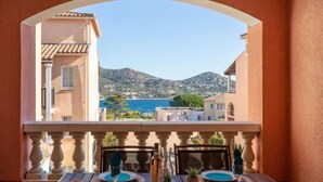 A balcony with a table set for dining, overlooking a coastal landscape with buildings and hills. - Le Village (C0 266) by Interhome (Agay Village Cap Esterel)