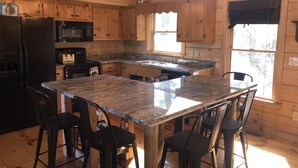 A wooden cabin kitchen with granite countertops, a central island with bar stools, a chandelier, and a large window. - Fantastic Mountain Views Plus Hot Tub and Fire Pit!! (Blue Ridge)