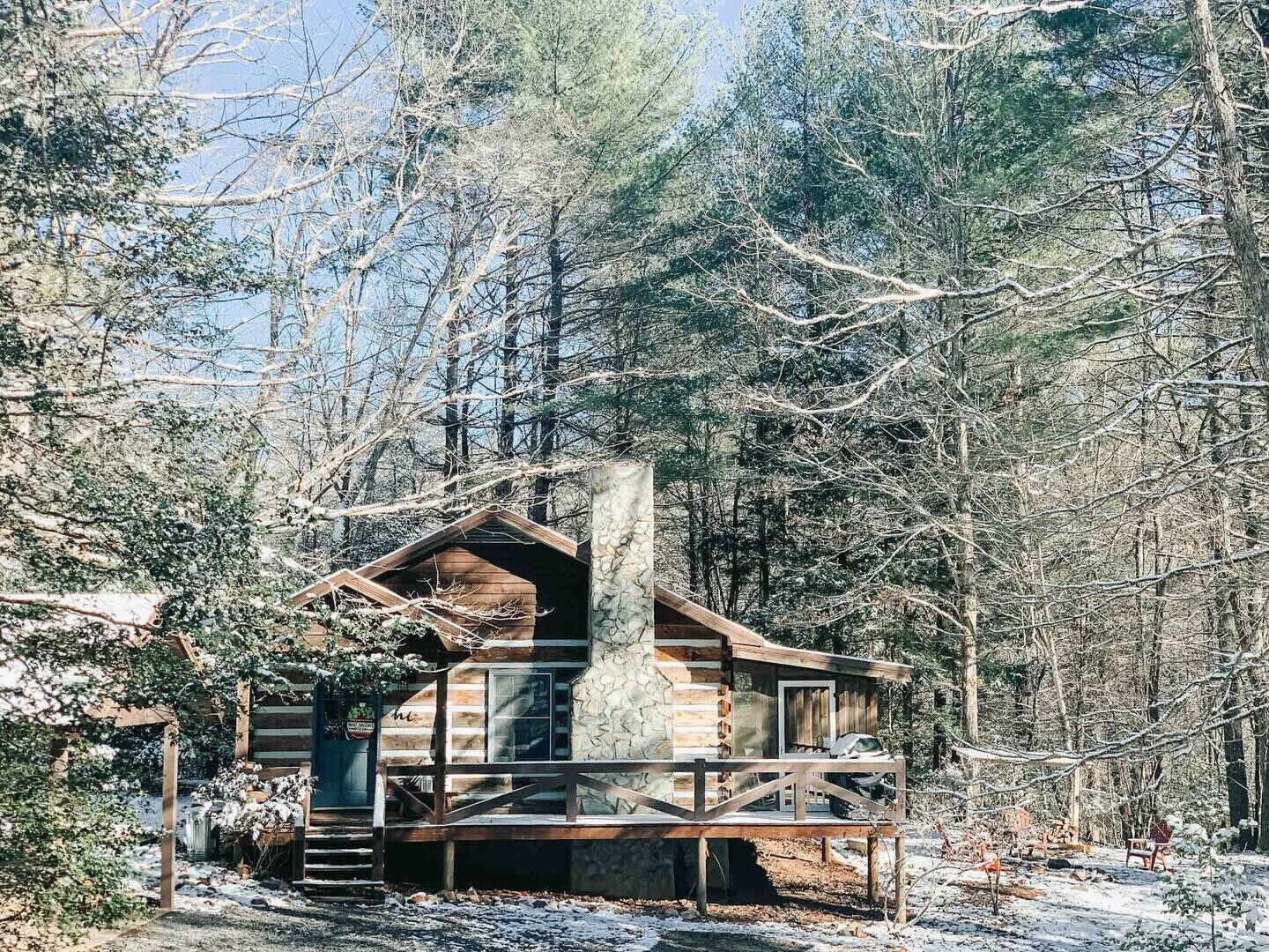 A log cabin with a stone chimney, surrounded by a wooden deck and snow-covered ground.