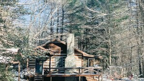 A log cabin with a stone chimney, surrounded by a wooden deck and snow-covered ground. - Cozy Cabin in woods with Hot Tub, Porch Swing Bed and Fire Pit in Aska Adventure (Blue Ridge)