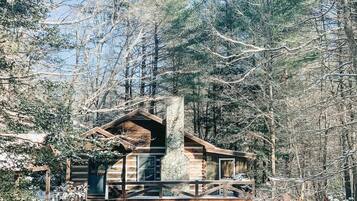 A log cabin with a stone chimney, surrounded by a wooden deck and snow-covered ground.