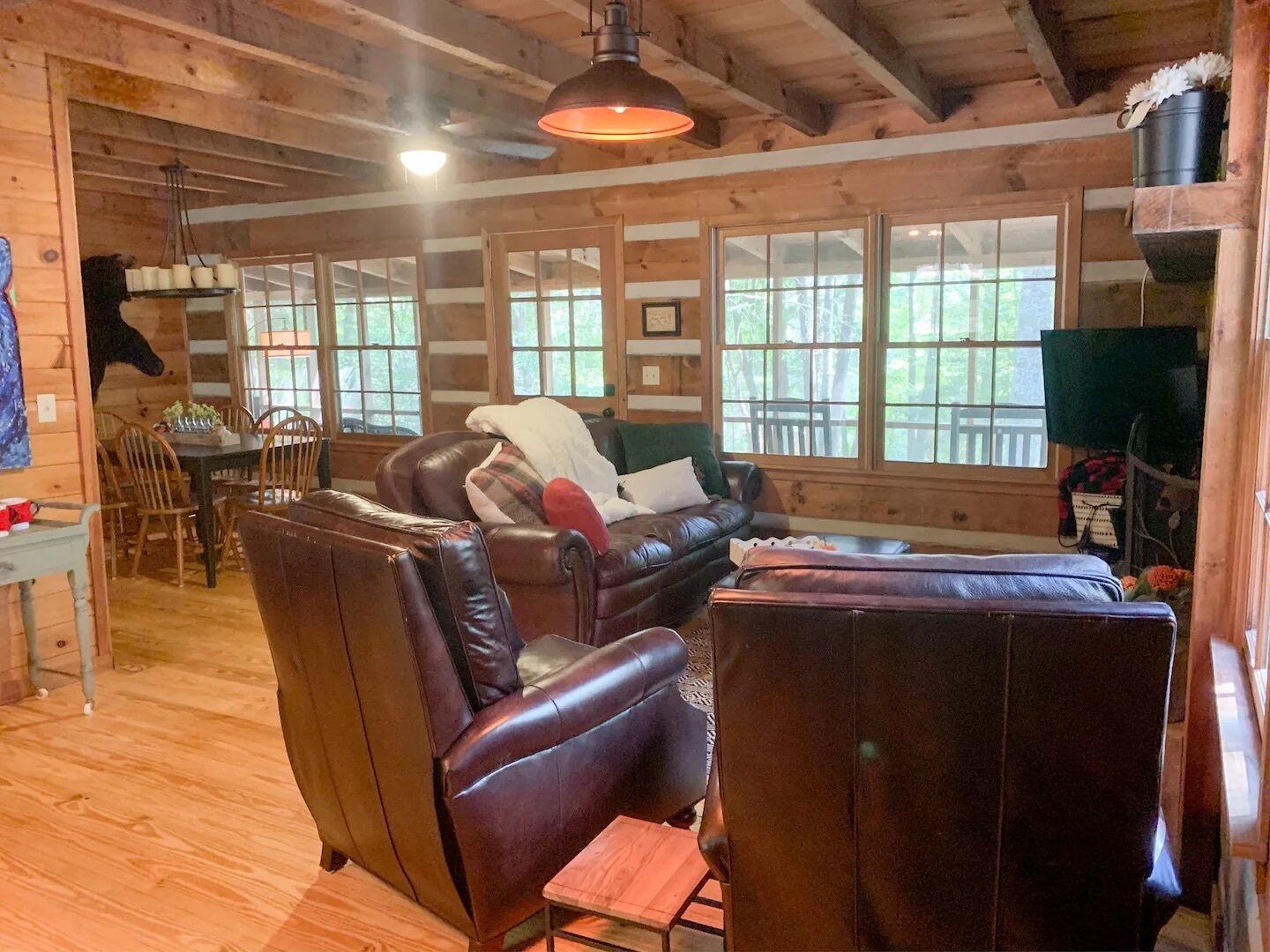 A cozy cabin interior with a leather sofa, a dining table with chairs, and large windows offering a view of the outdoors.