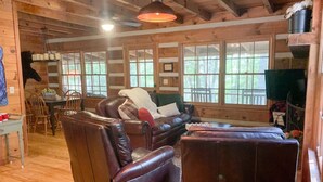 A cozy cabin interior with a leather sofa, a dining table with chairs, and large windows offering a view of the outdoors. - Cozy Cabin in woods with Hot Tub, Porch Swing Bed and Fire Pit in Aska Adventure (Blue Ridge)