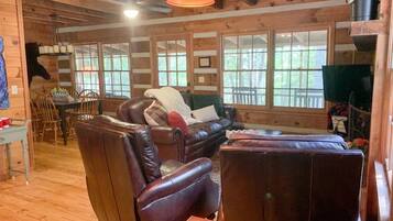 A cozy cabin interior with a leather sofa, a dining table with chairs, and large windows offering a view of the outdoors.