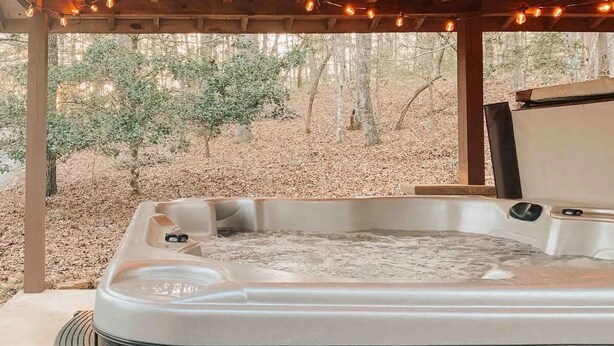A hot tub under a wooden pergola with string lights, surrounded by trees and a leaf-covered ground. - Cozy Cabin in woods with Hot Tub, Porch Swing Bed and Fire Pit in Aska Adventure (Blue Ridge)