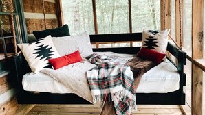 A wooden cabin with a rustic interior, featuring a wooden swing bed with plaid bedding and a forest view through large windows. - Cozy Cabin in woods with Hot Tub, Porch Swing Bed and Fire Pit in Aska Adventure (Blue Ridge)