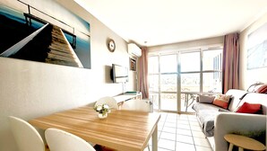 Un salon moderne avec une table à manger en bois, des chaises blanches, un canapé avec des coussins, un téléviseur fixé au mur et une grande fenêtre avec des rideaux.
