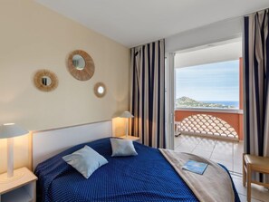 A hotel room with a bed, two bedside lamps, a view of the sea, and a balcony with a patterned railing. - Les Bastides (I4143) by Interhome (Agay Village Cap Esterel)