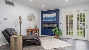 A modern living room with a leather chair, a wooden side table with books, a decorative wall piece, a large painting, and a potted plant. - Golden Racehorse MCM 3BR Slps 6 - 7 Min to Oaklawn (Hot Springs)