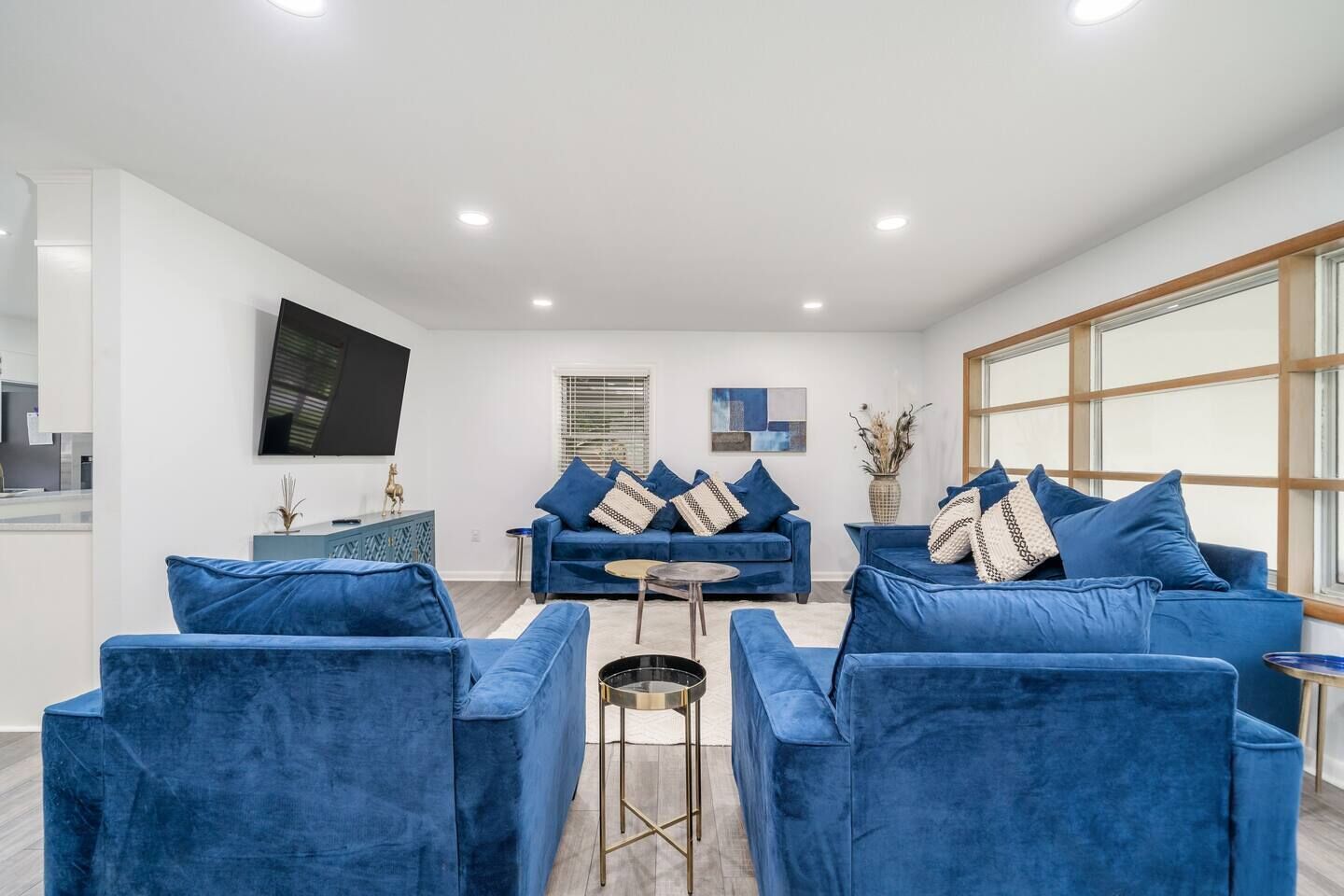 A modern living room with a blue sofa set, a flat-screen TV mounted on the wall, and a large window providing natural light.
