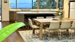 A modern kitchen with a large island, a dining area with a wooden table and chairs, and a view of the outdoors through large windows. - Hanging Garden On Mernda- Mountain view retreat  (Olinda)