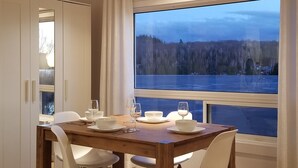 Ruokailutila, jossa on kahdelle katettu pöytä, suuri ikkuna, josta avautuu näkymä metsään ja vesistöön, sekä valkoiset tuolit.