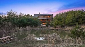 Eine hölzerne Hütte mit einem Balkon, umgeben von einem Teich und dichtem Baumbestand.