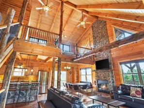 A spacious log cabin interior with a stone fireplace, a dining area, and a balcony. - Longview Lodge 5BR + loft, Fishing Pond, Hot Tub, Fire Pit, Close to everything! (Rockbridge)
