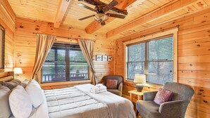 Ein Schlafzimmer einer Holzhütte mit einem Bett, zwei Sesseln, einem Deckenventilator und großen Fenstern mit Jalousien.