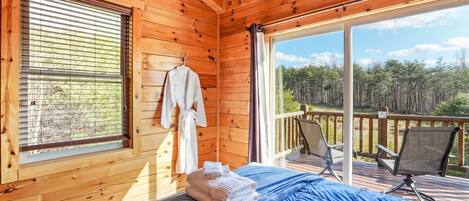 A wooden cabin room with a bed, a window with blinds, a balcony with chairs, and a view of a forest.