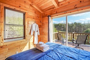 A wooden cabin room with a bed, a window with blinds, a balcony with chairs, and a view of a forest. - Longview Lodge 5BR + loft, Fishing Pond, Hot Tub, Fire Pit, Close to everything! (Rockbridge)