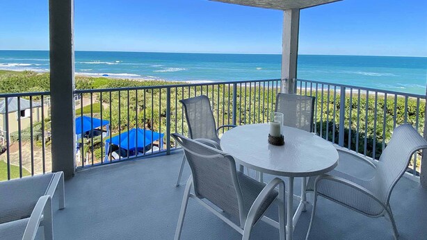A balcony with a view of the ocean, white chairs, a round table, and blue umbrellas. - The Sea Turtle at Spoonbill Resort Villa (Stuart)