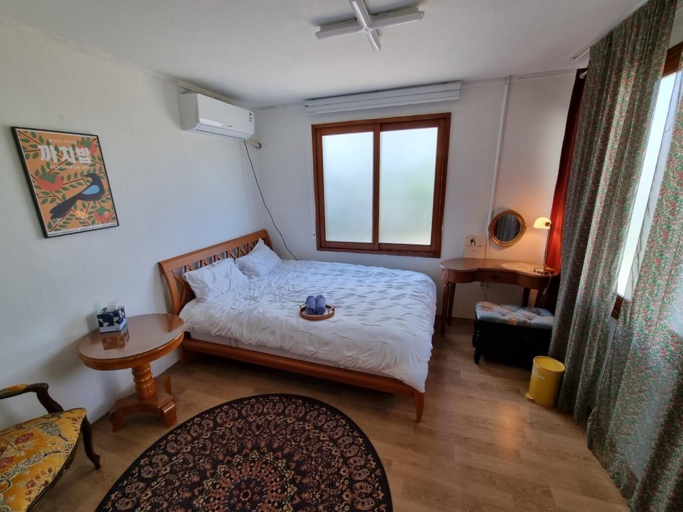 A bedroom with a bed, a bedside table, a chair, a desk, a mirror, a window, and an air conditioning unit.