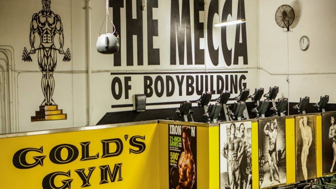 A gym interior with a large sign reading 'THE MECCA OF BODYBUILDING' and a display of bodybuilding magazines.