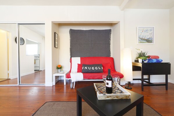 A living room with a red sofa, a black and white patterned tapestry, a wine bottle on a tray, and a small white table with a flower.