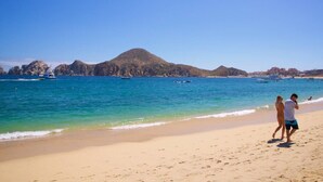 A beach with clear blue water, a sandy shore, and rocky hills in the background. - Casa Mismos w/pool |Walk to Medano Beach + Marina (Cabo San Lucas)