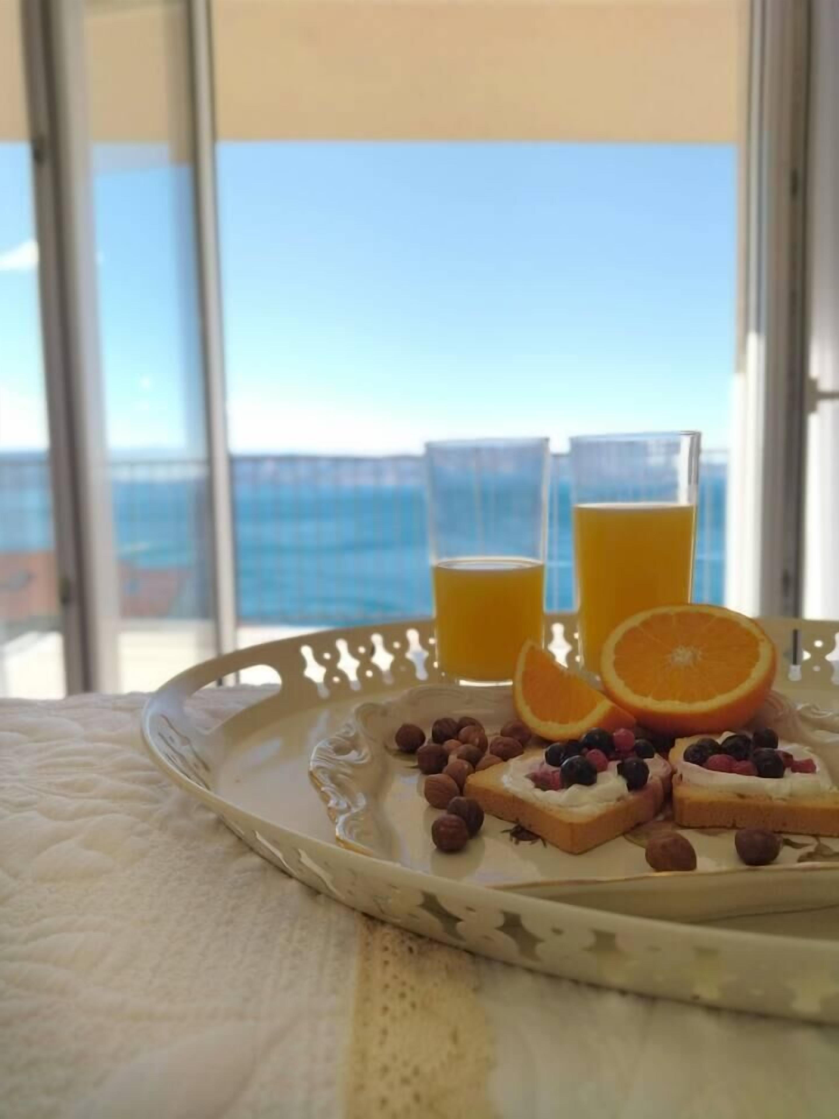 A breakfast tray with toast, fruit, and juice on a table by a window with a view of the sea.