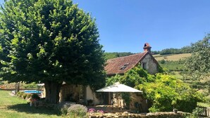 Une maison en pierre avec un toit de tuiles, un grand arbre au premier plan et un ciel bleu dégagé.