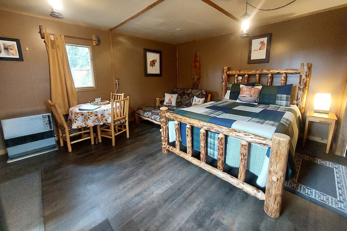 A rustic bedroom with a wooden bed frame, a small table with chairs, and a window with curtains.