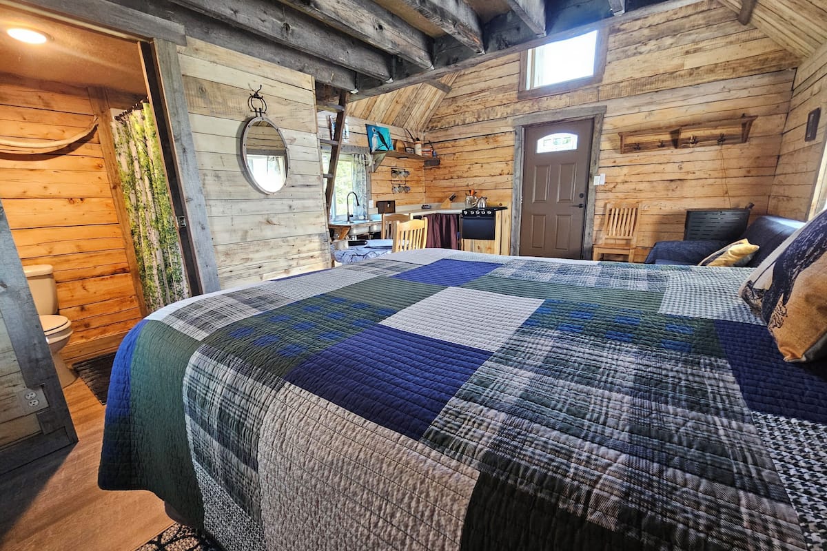 A log cabin bedroom with a large bed, a wooden ceiling, a small window, and a door.