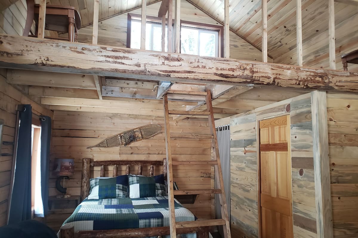 A rustic cabin interior with a loft bedroom, wooden beams, a ladder, and a bed with a plaid comforter.