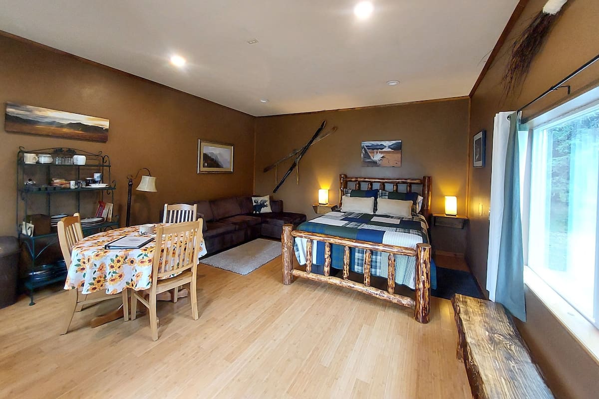 A cozy room with a wooden floor, a bed with a blue and white quilt, a wooden bench, a dining table with chairs, a bookshelf, and a window with curtains.