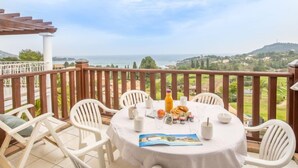 Une table ronde recouverte d’une nappe blanche, entourée de chaises en plastique blanches, est placée sur un balcon d’où l’on peut admirer un paysage magnifique sur la mer et les collines.