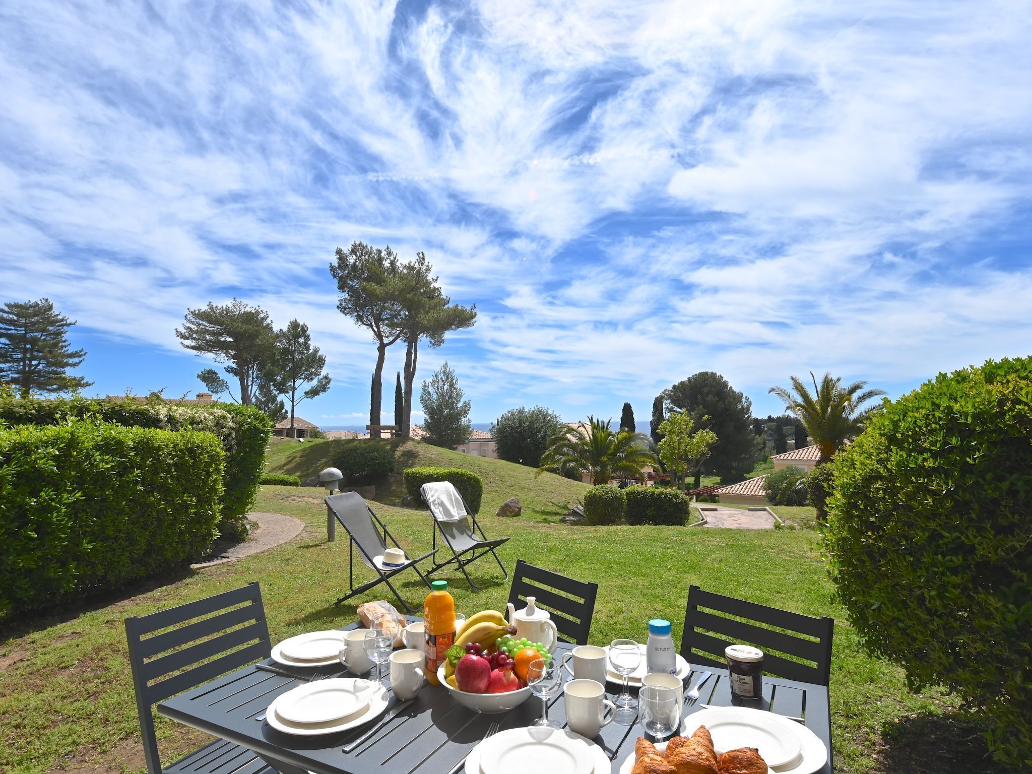 Une table à manger installée à l’extérieur, avec des assiettes, des tasses et un saladier rempli de fruits, entourée de verdure et d’arbres, sous un ciel dégagé.
