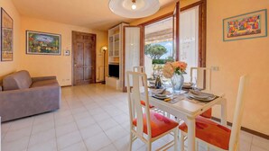 Een eetgedeelte met een tafel, stoelen, een vaas met bloemen en een bank in een kamer met betegelde vloer en een plafondlamp.