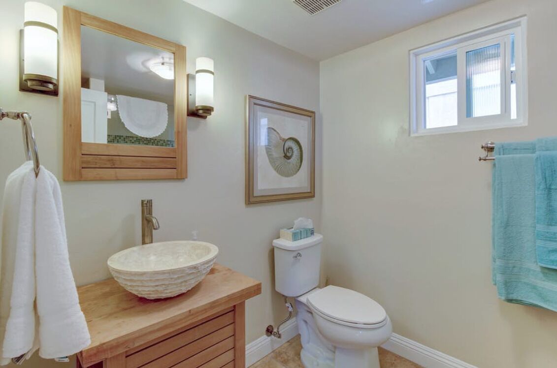 A bathroom with a wooden vanity, a bowl sink, a toilet, a framed artwork, and a window.
