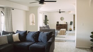 A modern living room with a dark-colored sofa, a ceiling fan, and a view into the dining area. - The Desert Sunset: 4 BDR, 5 Beds, Fast Wifi, Large TV, Large private pool, (Mesa)