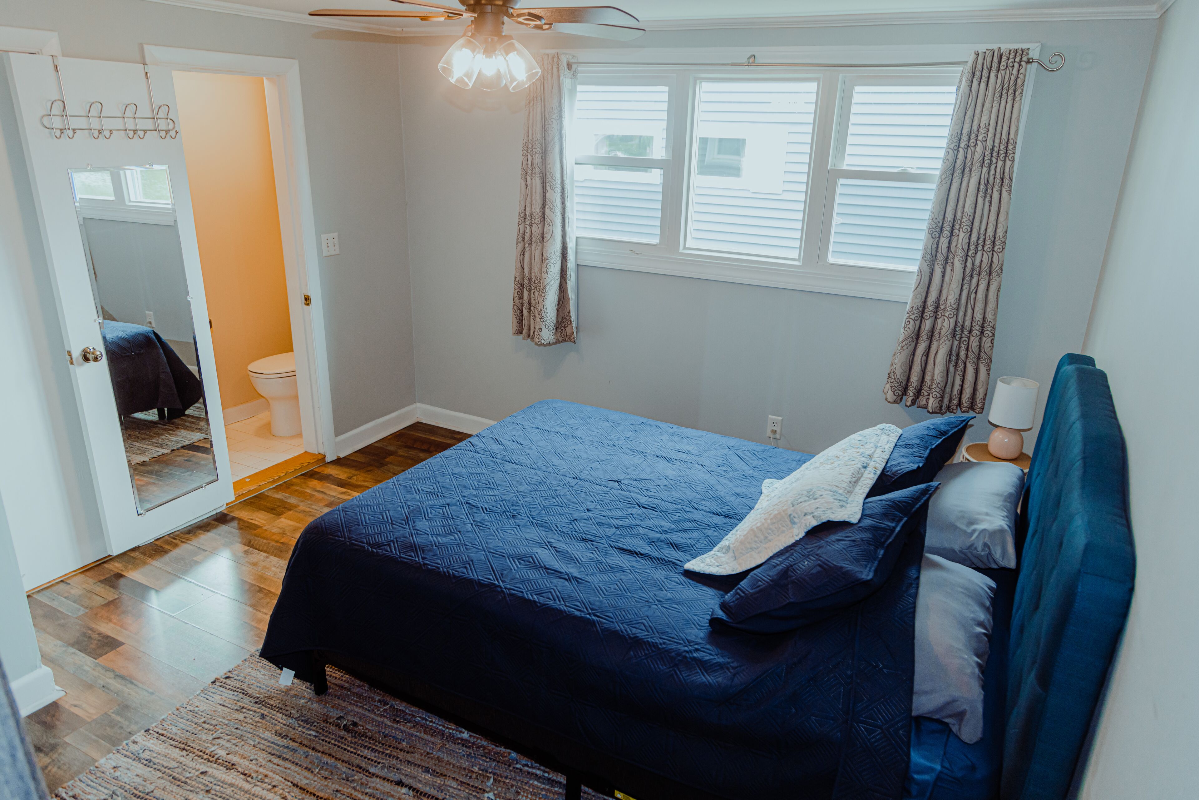 Un dormitorio con cama, ventilador de techo, ventana con cortinas y puerta que da a un baño.