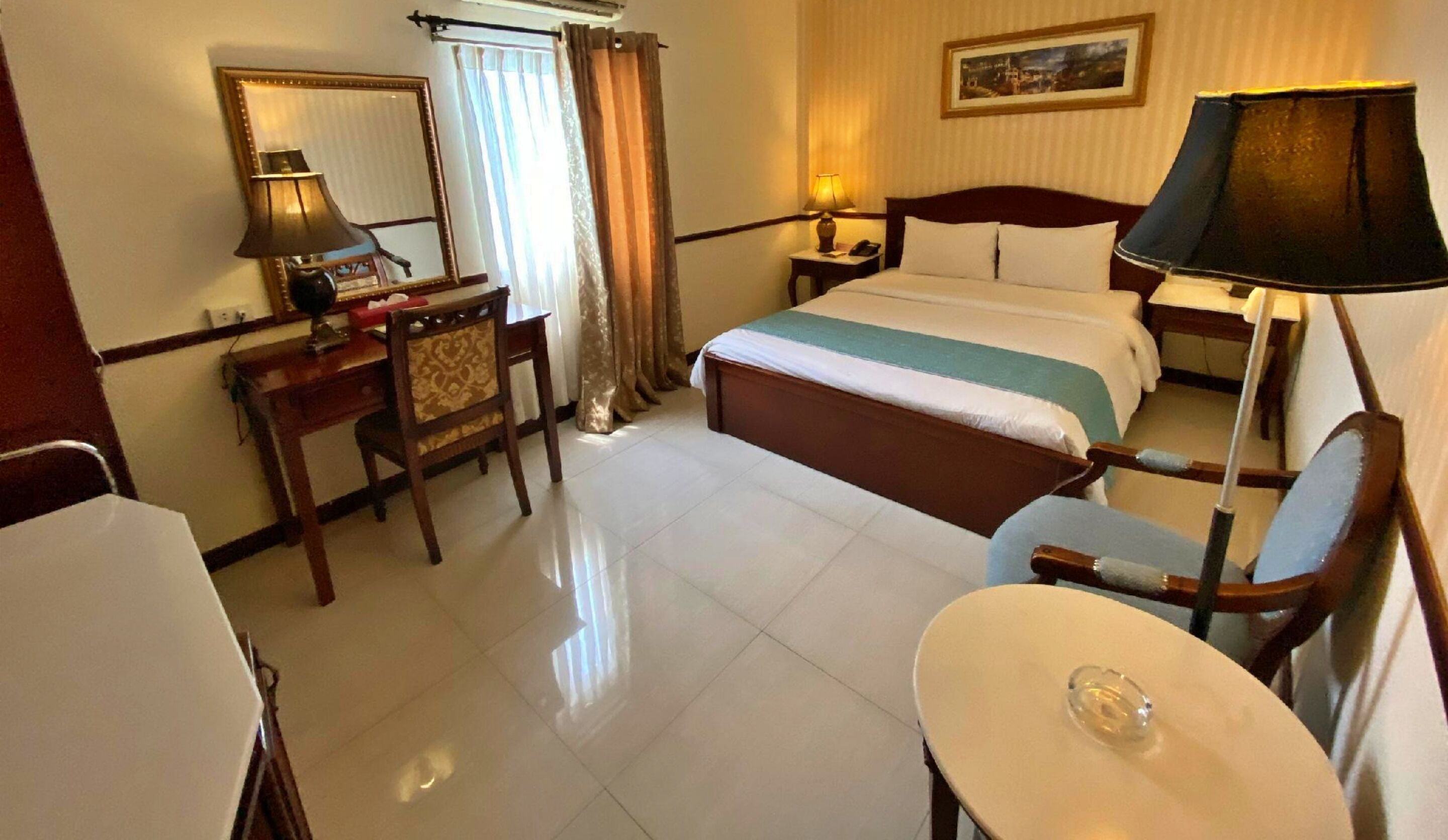 A hotel room with a bed, bedside table, lamp, chair, desk, and a small table with a glass object.