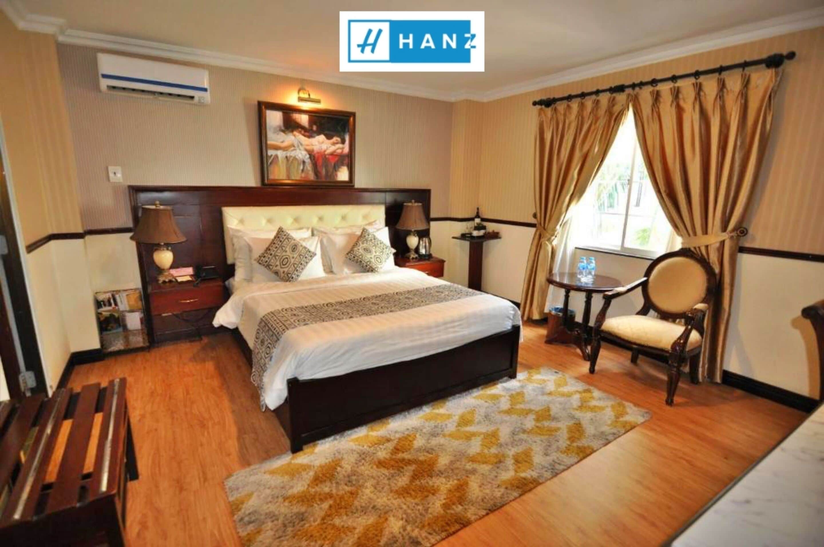 Ein Hotelzimmer mit einem großen Bett, einer hölzernen Bank, einem kleinen Tisch mit Stuhl und einem Fenster mit Vorhängen.