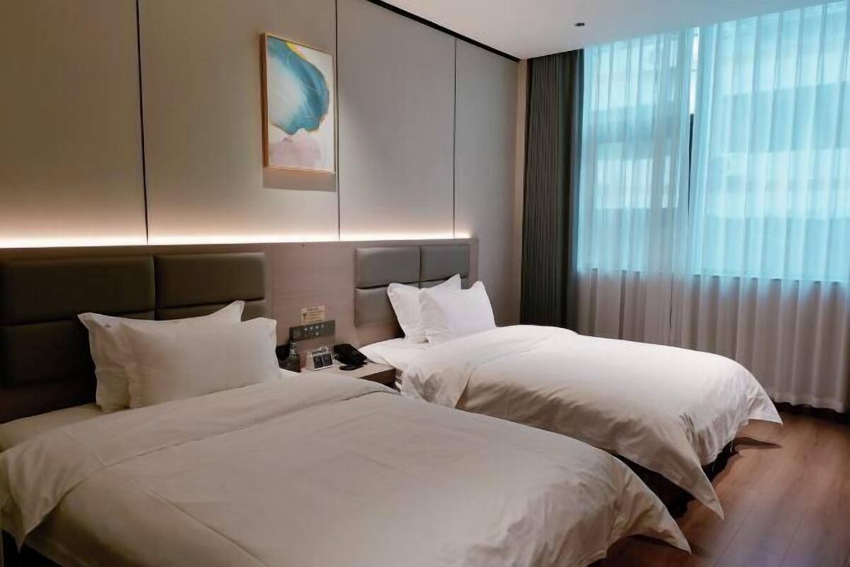Habitación de hotel con dos camas, una mesita de noche con lámpara, un cuadro en la pared y un ventanal con cortinas.