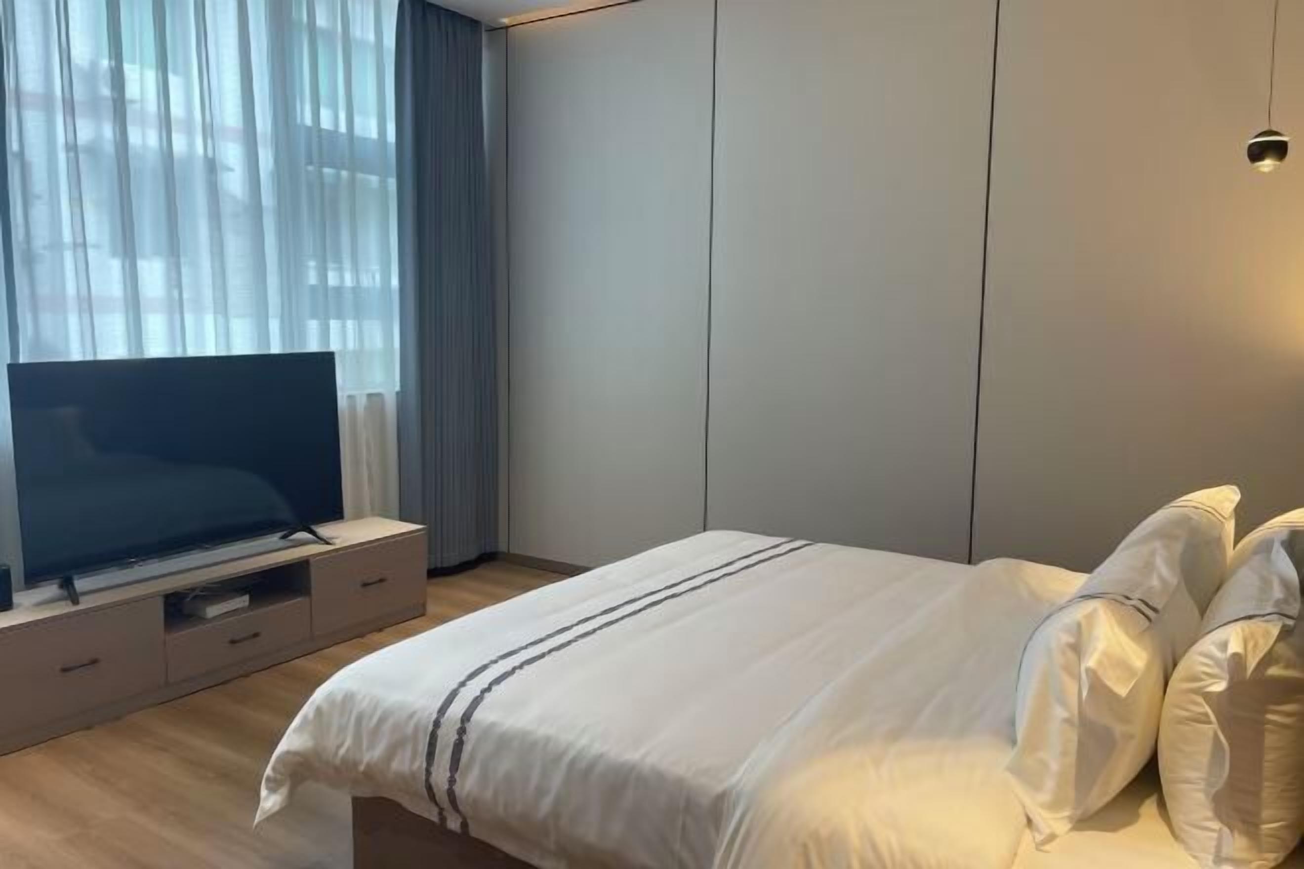 A modern hotel room with a large bed, a flat-screen TV on a wooden console, and a large window with sheer curtains.